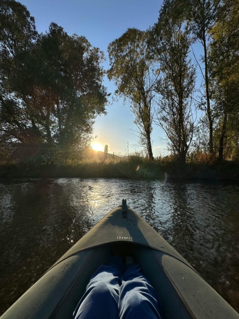 Vooraanzicht vanuit een donkergrijze packraft op een smalle, stille waterweg in Gaasterland. De bomen langs de oever staan in herfstkleuren en het zonlicht schijnt laag door de takken. De sfeer is rustig en avontuurlijk. Ideaal voor wie packraften in Friesland wil ontdekken.